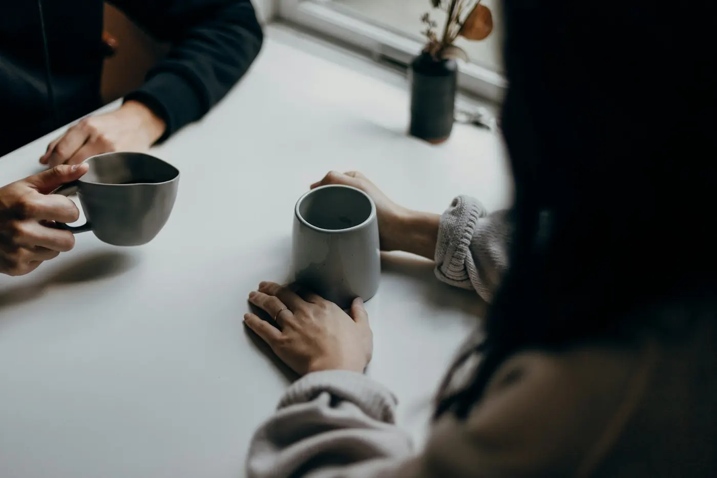 Two people talking over mugs at a table, hands visible
